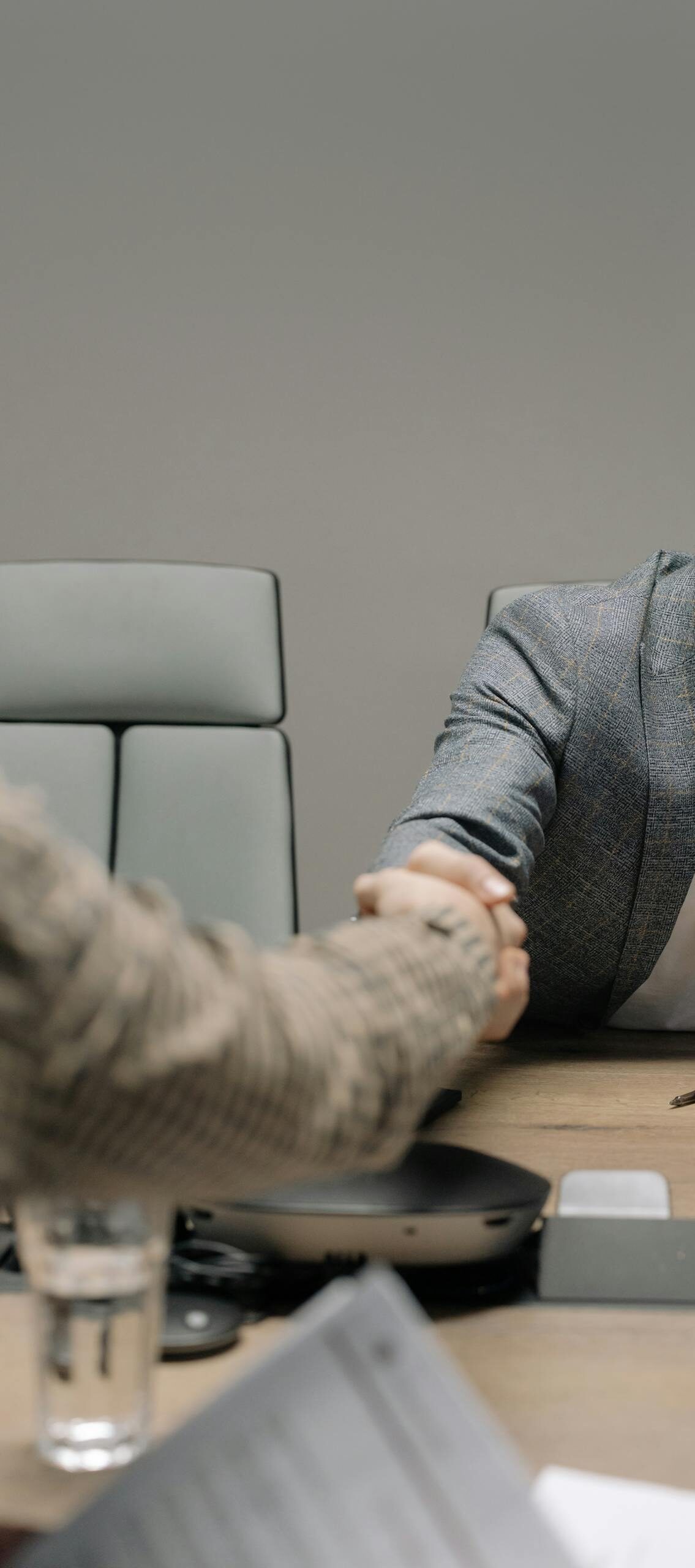 Two businessmen in a modern office shaking hands across a table. Professional setting.